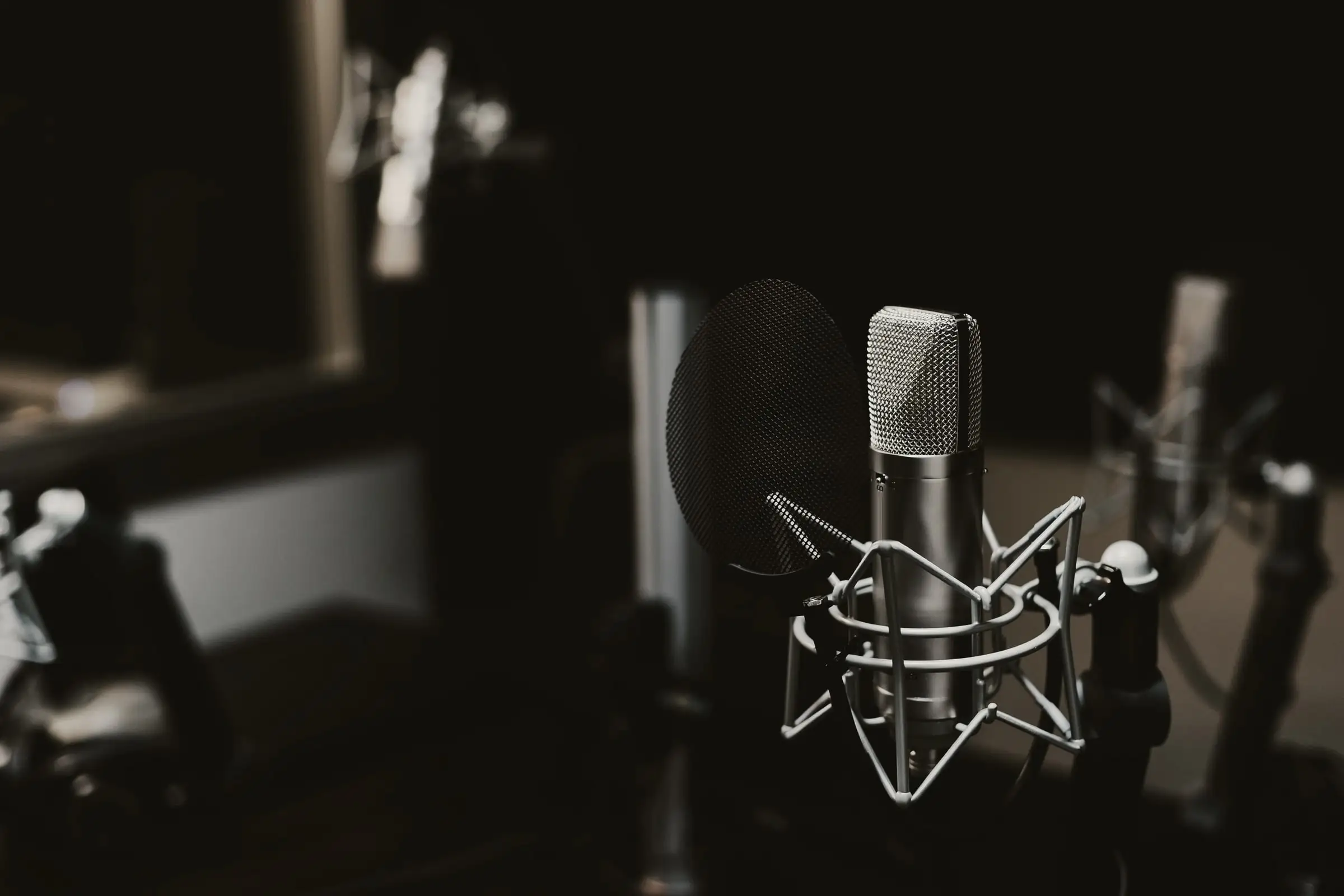 Silver condenser microphone in a shock mount behind a pop filter, dark recording studio, blurred gear behind