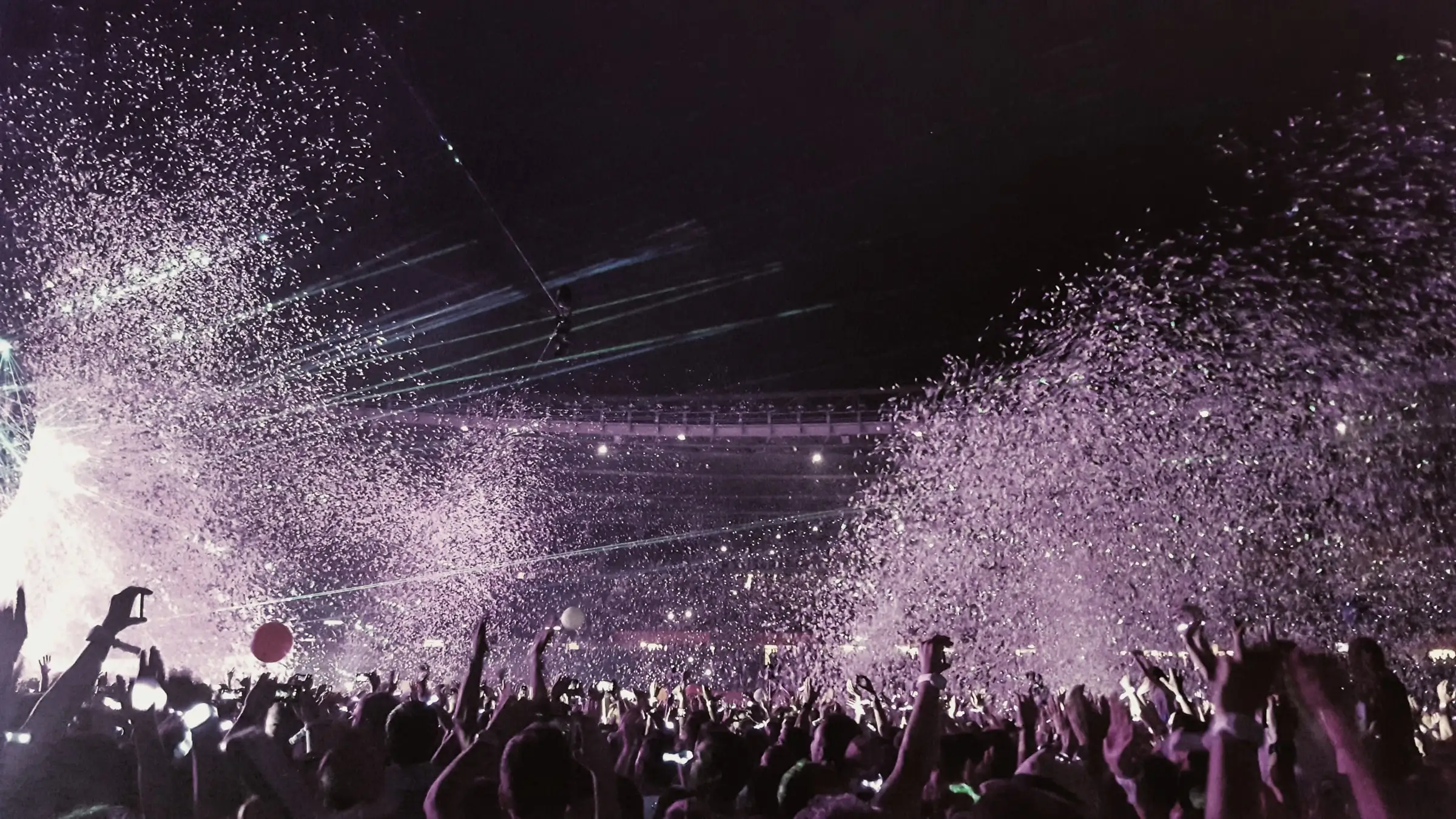 Stadium crowd from behind with raised hands and phone lights under purple confetti and stage light